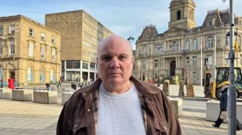 Gemma Dillon/BBC A bald man wearing a grey top and brown jacket is facing the camera. 
Behind him is Dewsbury Town Hall a stone building, and a boarded up building. 