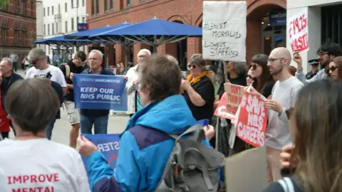 A group of people are holding banners with protest messages including 'Save NHS careers', 'Greater Manchester/keep our NHS public' and 'highest mortality/lowest staffing'.