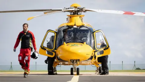 An air ambulance crewman, dressed in a red and black flying suit and carrying a white helmet, walks away from a yellow helicopter, which is parked on a runway and facing the camera. The doors of the helicopter are open and two other crew members can be seen behind them.