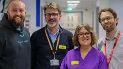 Royal United Hospital Bath Rugby player Tom Dunn is on the far right wearing a navy blue Bath Rugby hooded jumper. He stands next to Andrew Hollowood, who wears a naby blue fleece, blue shirt and a staff lanyard. Margi Jenkins is on his right wearing purple hospital scrubs and Dr William Headdon is on the far right wearing a white shirt and red lanyard. They are all standing in a hospital corridoor.