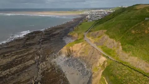 Getty Images A green hill with a coastal path cutting through it is seen next to the rocks and the sea. There is a town in the distance.