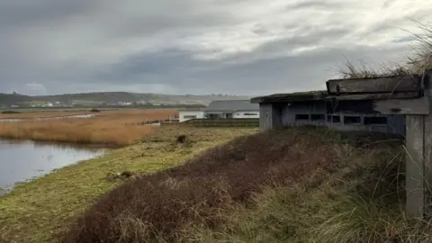 A natural brown looking building blending in with bushes and grassland with a large expanse of water to the left of it. Further afield are hills and houses with dark grey skies.