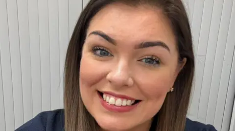 Clare has long dark hair and is smiling at the camera. She has blue eyes and is wearing pink lipstick. She is standing in front of white blinds.