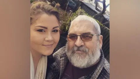 Sophie Leadsham A young woman with brown hair poses next to her father, an older man with glasses and a beard, both smiling at the camera