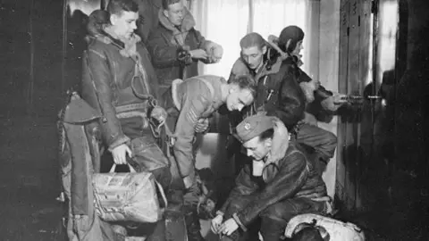 IWM Duxford A black and white image showing Lancaster pilots preparing in the crew room at RAF Duxford during World War Two. There are six men pulling on equipment and uniforms, one is opening a locker on the right. On either side are metal lockers. Behind them is a window. 