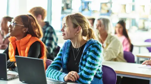 A female student with blonde hair and wearing a blue and green striped cardigan sitting at a desk in a classroom with a laptop. There are other students sitting on her left and behind her.