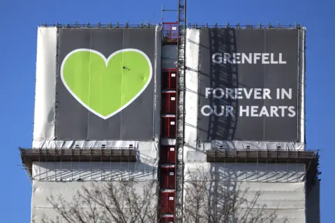 Image shows a close up of the top of Grenfell Tower with the message 'forever in our hearts' and a green heart.