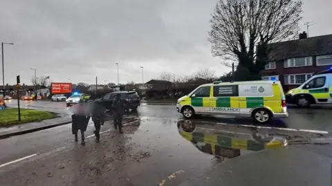 Steve O'Leary Ambulances and police cars in the rain in King George's Avenue, Harwich, with schoolchildren walking past