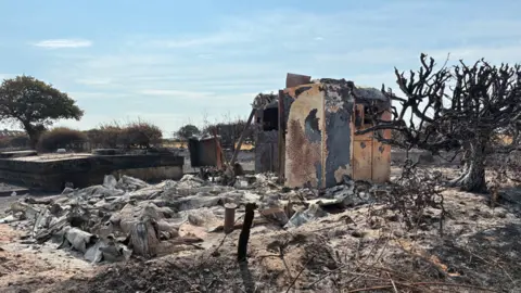 Remains of a small building surrounded by burnt grey remains on charred ground.