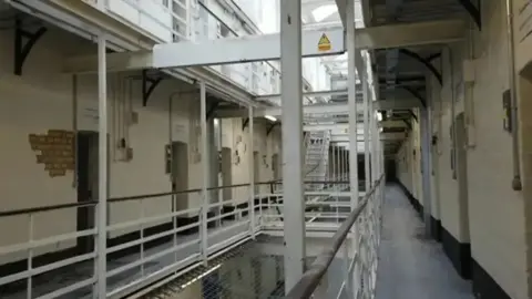 A prison wing inside a Victorian prison. Cell doors are visible on either side of a long corridor with stairways and a netted gap in the middle. Part of the walls show exposed brickwork. 