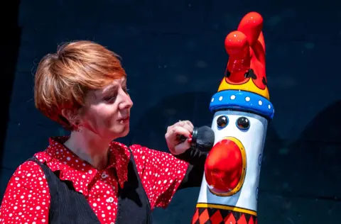 A woman polishes a Feathers McGraw statue at the unveiling of the Gromit Unleashed 3 trail in Bristol. The statue is painted in bright colours and the woman is wearing a red and white shirt with a black cardigan over the top of it