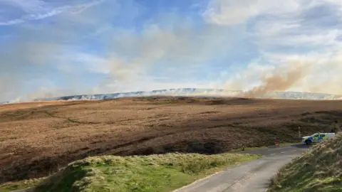 A line of fire and smoke stretching across brown moorland with a police van in white, blue and florescent yellow livery parked at a road junction