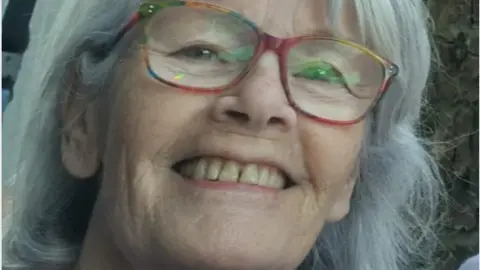 A close-up of the late Bernadette (Bernie) Cranley.  She has shoulder-lengthy grey hair and is wearing glasses with coloured rims. 
