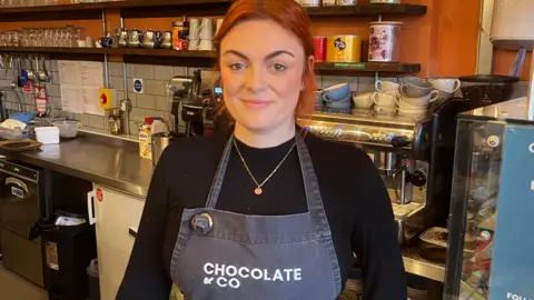 BBC/RICHARD EDWARDS A woman is standing in front of a coffee machine, shelves and a counter. She has red hair tied back, dark eyes and an eyebrow piercing. She is wearing a long sleeved black top and an apron with Chocolate & Co written on the front. Behind her, cups are piled on top of the silver coffee machine. Tubs, glasses and silver pots are neatly stacked on a long shelf.