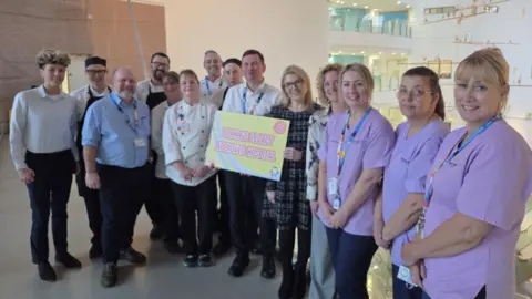 Alder Hey NHS Trust About a dozen Alder Hey workers smile as they line up to pose for a picture. A man and woman in the centre hold up a placard saying it's a hospital approved by Sophie's Legacy.