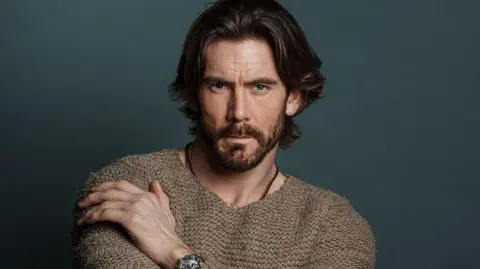 A headshot of James Wilkinson in a brown knitted jumper. He is posing while sat down in front of a dark green background. He has long brown hair and a dark beard.