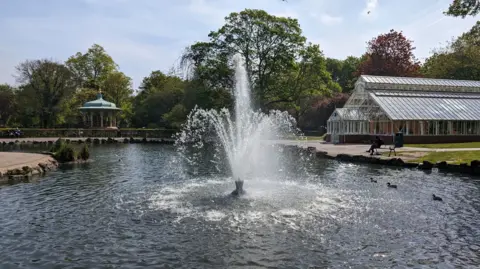 View of Pearson Park pond. It is curved and surrounded by pathways with a bandstand in the background.