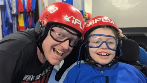 Dave Clarke Dave, left, and Addy, pictured in helmets and goggles ready for an indoor skydiving experience. Both are smiling widely at the camera. 