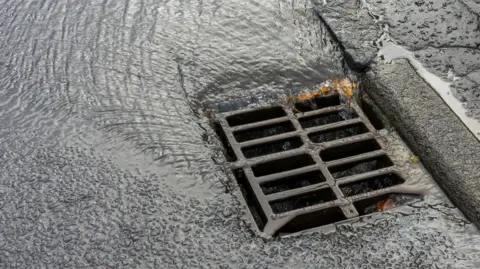 Getty Images Close-up of a wet road gully with rainwater flowing into it. The surrounding road surface and kerb are slick with water after rainfall.