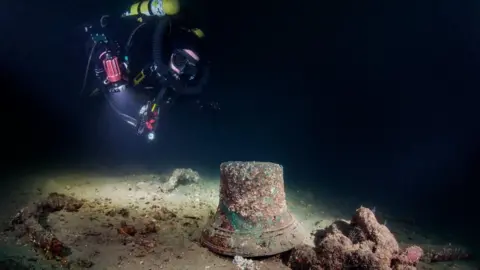 An underwater picture with a diver approaching a bell, with marine life on it from above.