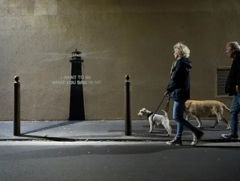 Banksy / Instagram A photo of the Banksy piece of art with two dogwalkers walking past on the street. A metal bollard is in the foreground on the pavement, while a painting of a lighthouse is on the wall superimposed with a quote "I want to be what you saw in me".