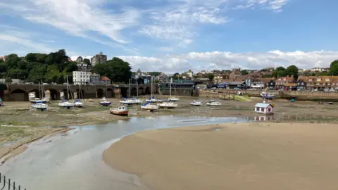 Low tide at Folkestone harbour. Light brown sand in the foreground, houses behind with blue and cloudy sky above.