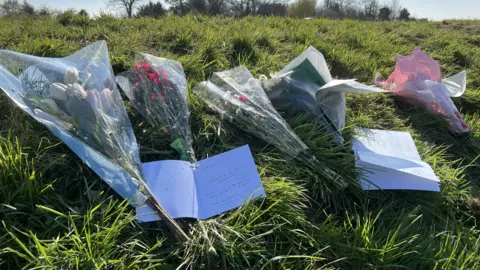 Mousumi Bakshi/BBC Flowers and cards are lying on a river bank. There are five bouquets and two square cards with written words on them.