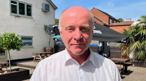 A bald man wearing a white shirt is looking directly at the camera. He is standing outside a white-painted pub with wooden picnic tables and large umbrellas and small trees outside.