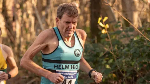 David Griffin David Griffin is running wearing a sky blue vest with Helm Hill and the number 44 on the front. He is a man in his 60s with brown hair and a determined expression. Behind him is a canopy of trees and a part of another runner, in yellow and green, is just in shot behind him.