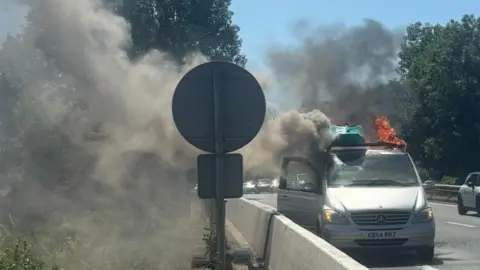 A silver van by the side of a dual carriageway with a kayak on the roof, flames and smoke billowing from it.