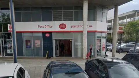 A post office on Midsummer Boulevard, Milton Keynes. It is a ground floor corner unit of a bigger building. Above the floor length windows is a sign saying post office is red letters. There is also a Post Office logo above the door saying post office in white letters on a red oval. A woman is standing to the left of the door. 