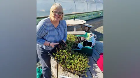 Woman with glasses and grey hair wearing a striped blue top and smiling by table with lots of plants on it. 