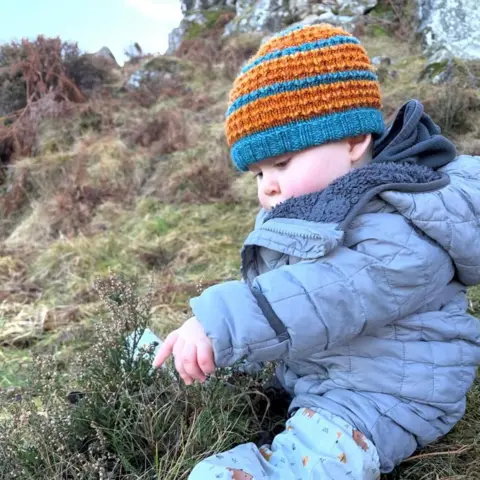 Morag Skelton Hamish sitting on a hillside wearing a blue jacket and a striped hat.