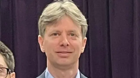 Sue Dougan/BBC Head and shoulders image of Brett Mickleburgh, who has light hair parted in the middle. He is wearing a grey suit and blue shirt and is standing in front of a purple curtain.