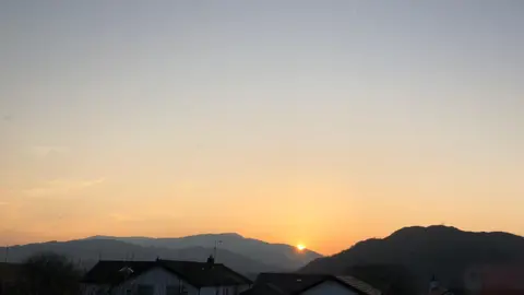 BBC Weatherwatchers Sun coming up behind hills with houses in the foreground. The sky is an orange colour. In the distance are the peaks of large hills.