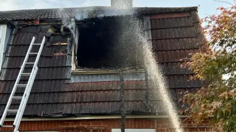 ECFRS The roof of a house where the force of a fire has blown out a dormer window. A ladder is propped against the roof and a jet of water from a fire hose is being sprayed through the window void and into the building.