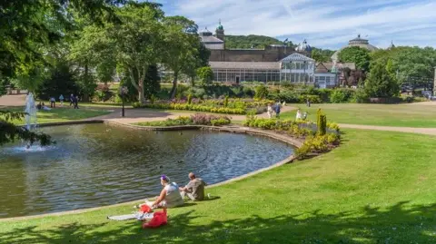 Getty Images A couple sits on a lawn next to lake in the middle of a large park on a sunny day. Behind them is a large pavilion, orangery and dome-roofed building.
