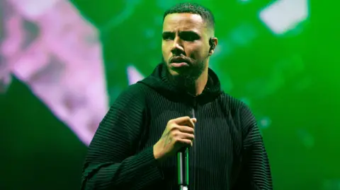 AJ Tracey performs on stage at Radio 1's Big Weekend. He wears a zip-up black hoodie and holds a microphone in his right hand, which has an M tattooed on the back. AJ has short dark hair and a beard and looks to the left of the camera. The staging behind him is lit green. 