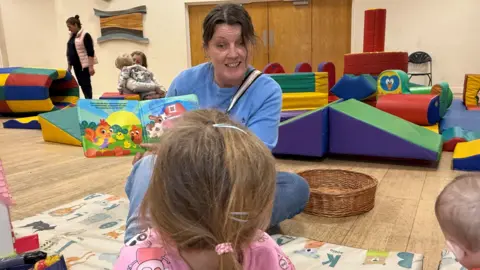 Steve Hubbard/BBC Mum Tanya reading to her daughter in a playgroup setting with soft toys in the background. Tanya, wearing a light blue top and blue jeans, is sitting cross-legged and holding a book up to her daughter who has her back to the camera.      