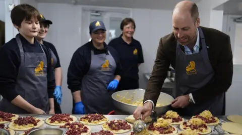 PA Media Prince William is holding a pasty. He is wearing a brown jacket and a blue apron.