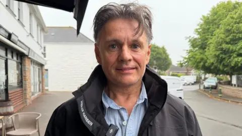 Carl Parslow is wearing a black jacket and a blue shirt with white stripes. He is pictured in St John's village.