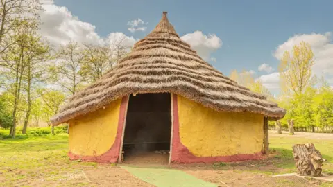 Flag Fen A roundhouse replica with a thatched roof. The walls are made of mud/clay with a wide door-shaped entrance. The exterior wall is painted saffron yellow. There are trees behind the roundhouse.