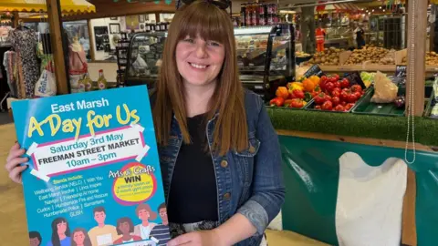 Local councillor Nicola Aisthorpe standing in front of a market stall holding a poster promoting the event. She has long brown hair and is wearing a black top and denim jacket.