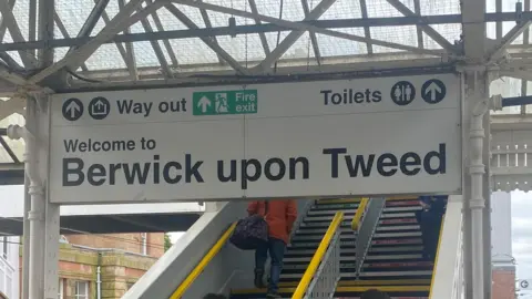 A sign saying "Welcome to Berwick upon Tweed" at the station, above some stairs which a passenger is climbing up.