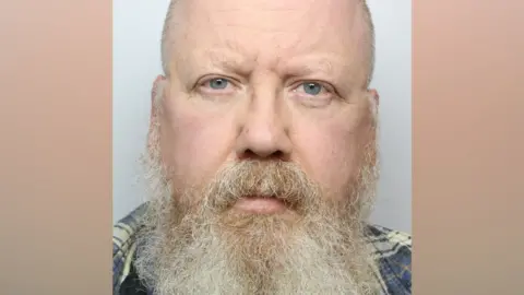 A man with a bald head and long grey beard. He is wearing a blue checked shirt and standing against a grey background