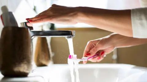 A pink toothbrush being held under a tap in a bathroom. A woman's hand is pressing down on the tap. 