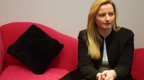 Alamy A young Michelle Mone on a bright red sofa. She is wearing a black jacket, white blouse, and looks very serious. There is a black cushion.