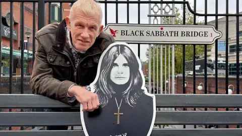 Westside BID Brian Hughes next to a cut out drawing of Ozzy Osborne on the Black Sabbath bench. Mr Hughes is wearing a brown jacket and has a hand on the bench. Behind him a sign says 'Black Sabbath Bridge'. 