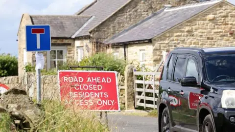LDRS A village lane with a one-storey stone cottage, gate and partial view of a car, a cul-de-sac sign and another stating that the road ahead is closed with residents access only.