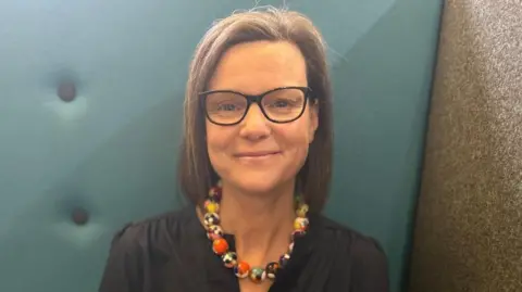 A headshot of a woman who is sitting on a blue sofa. She is wearing a black blouse and a chunky necklace that is made of yellow and orange marbles. She has brown hair that is bobbed and is wearing black glasses.